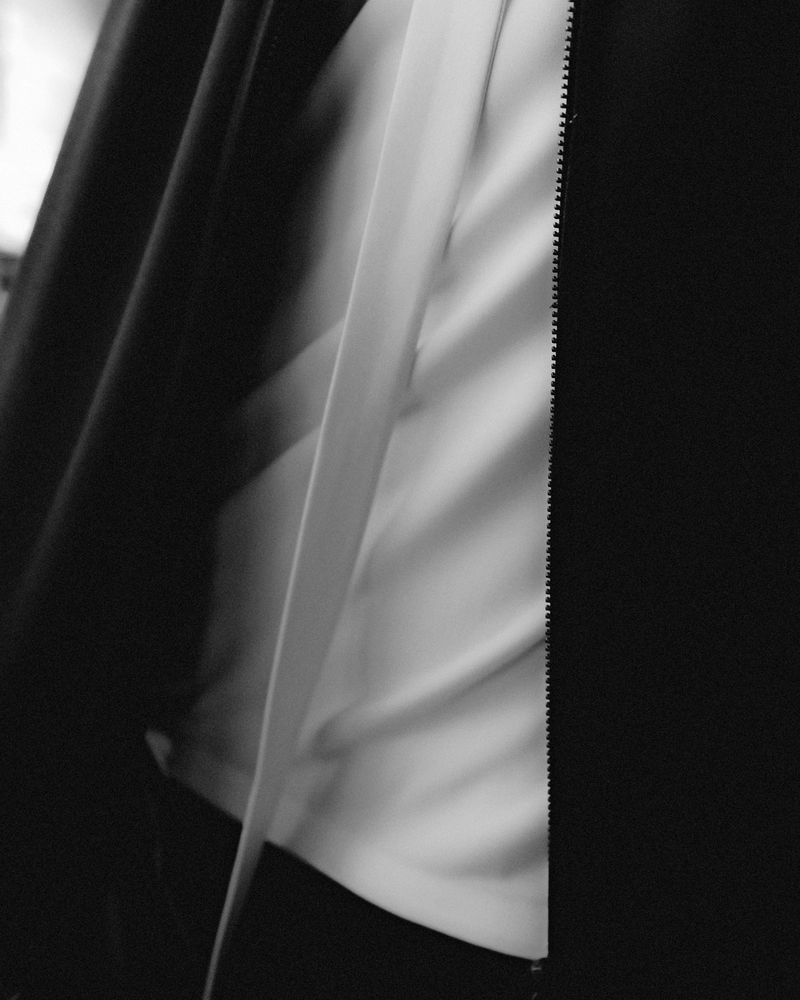 Monochrome image of a black coat partially open, revealing a white shirt underneath with a zipper on the side, creating a textured effect.