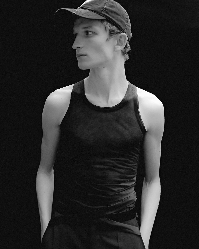 Black and white portrait of an adult male with dark hair, wearing a tank top and a baseball cap, looking off to the side.