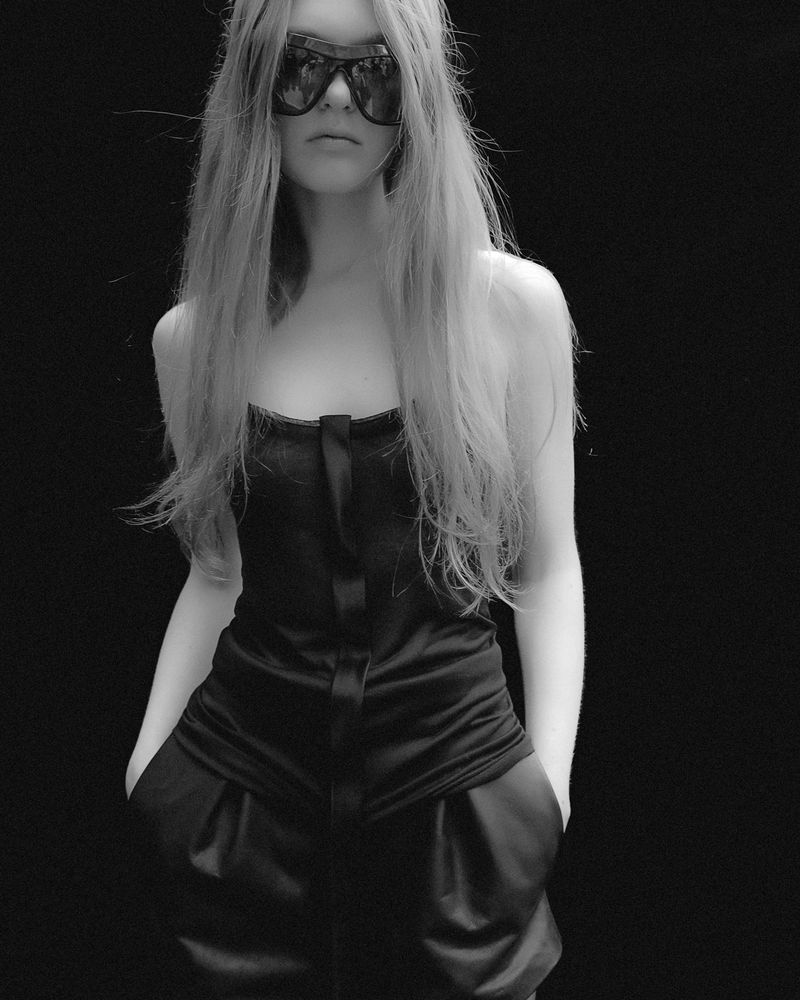 Black and white portrait of a slender young woman with long blonde hair wearing dark sunglasses and a dark satin romper suit.
