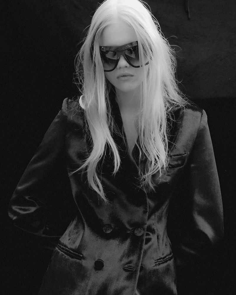 Black and white portrait of a young woman with long white-blonde hair wearing sunglasses and a dark double-breasted jacket, dark background.