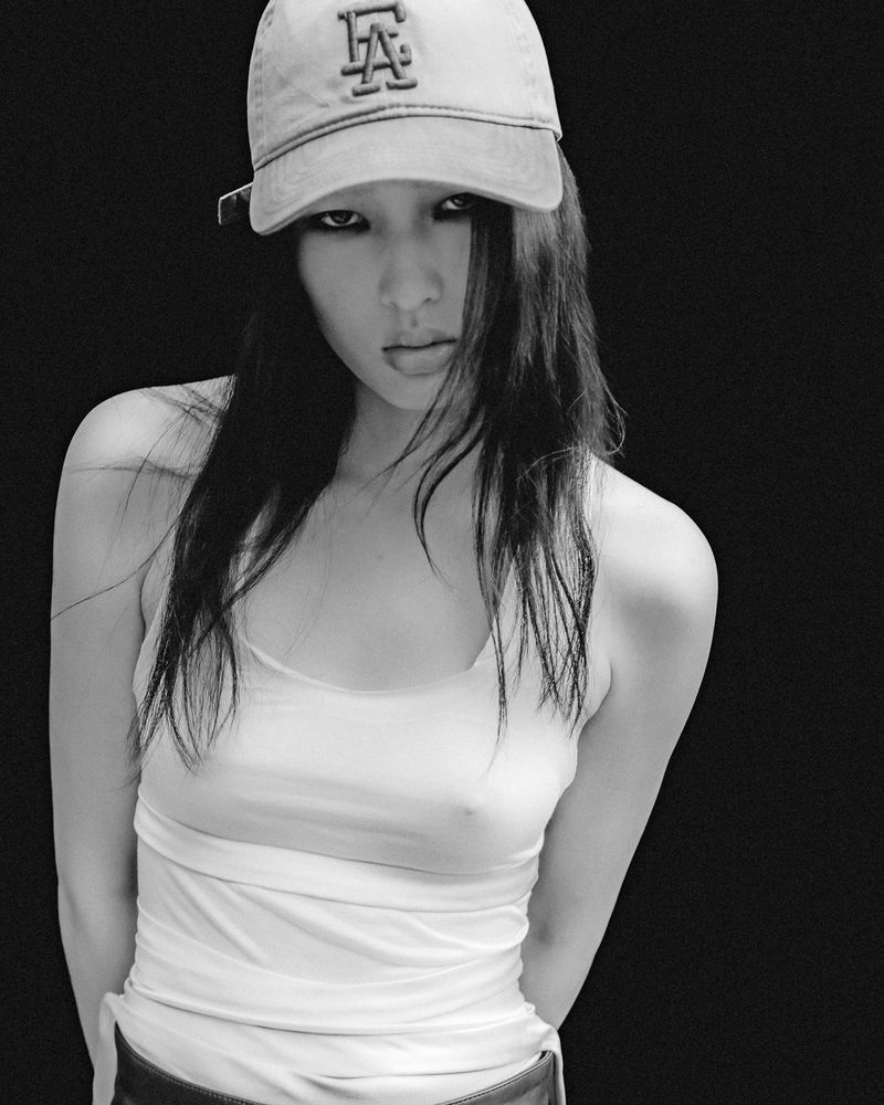 Model wearing a baseball cap and white tank top against a dark background in a black and white portrait.