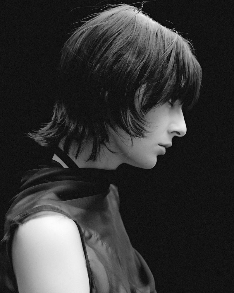 Monochrome portrait of a model in profile wearing dark garments against a solid black background.