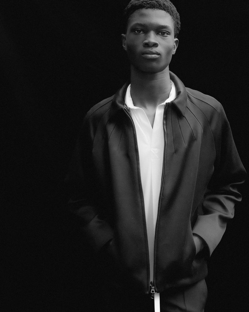 Black and white portrait of a young black man wearing a dark jacket over a white polo shirt against a plain dark background.