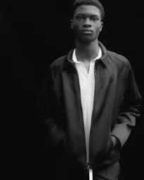 Black and white portrait of a young black man wearing a dark jacket over a white polo shirt against a plain dark background.