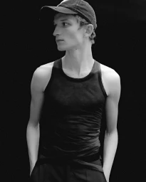 Monochrome portrait of a young male model wearing a dark tank top and a baseball cap against a black background.