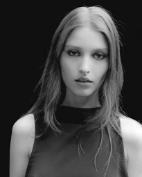 Close-up portrait of a model with dark eye makeup and long hair wearing a dark sleeveless top against a black background