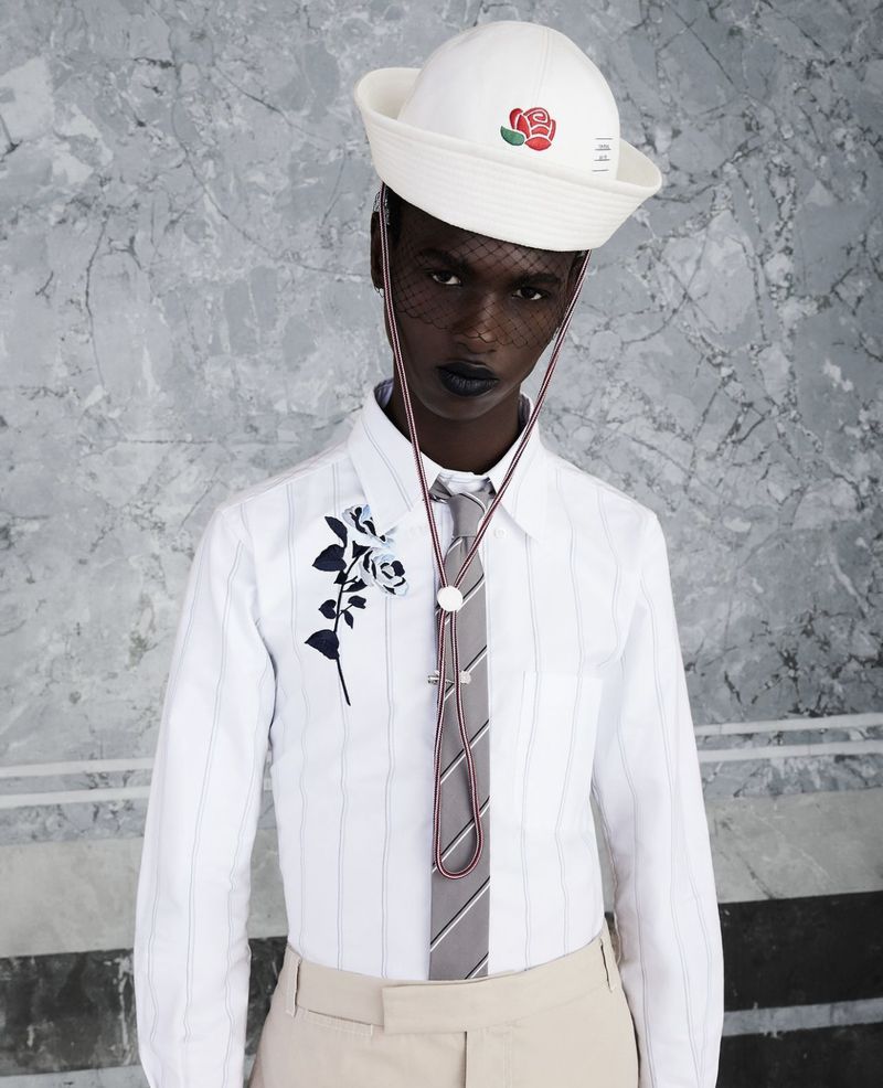 Person poses in pinstripe shirt, tie, and sailor hat with rose emblem in front of marble wall. Dark lipstick fashion portrait.