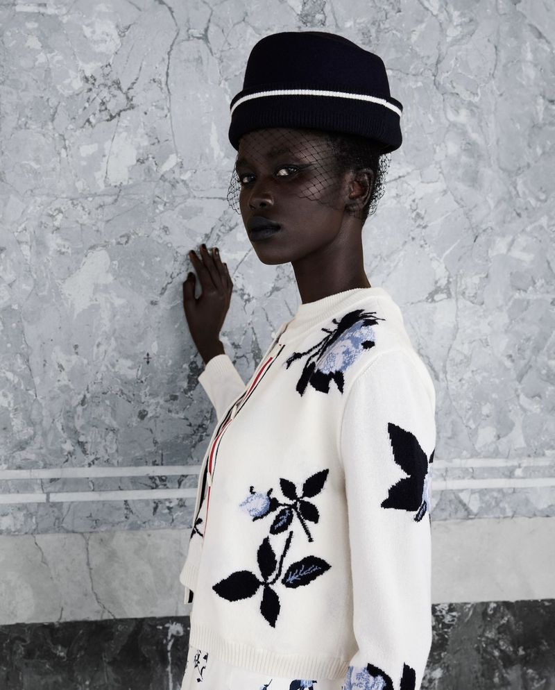Elegant woman in a black hat and floral sweater poses against a marble wall, showcasing fashion and style with a modern aesthetic.