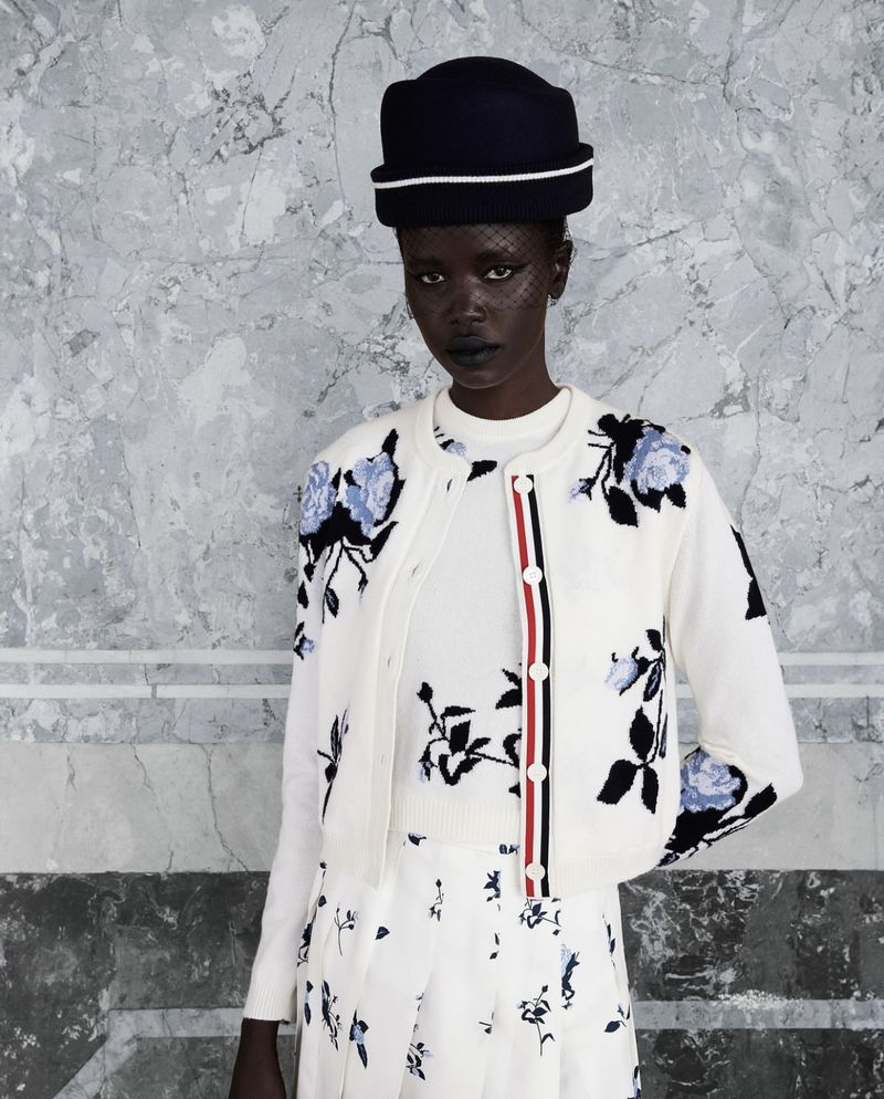 Fashion shot of a single Black woman wearing a floral-print sweater, hat, and skirt standing against a marbled background in a studio.