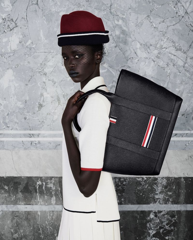 Fashionable woman with dark skin and hat poses with a black tote bag in front of a grey marble wall, showcasing modern style.
