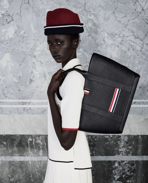 Fashionable woman with dark skin and hat poses with a black tote bag in front of a grey marble wall, showcasing modern style.