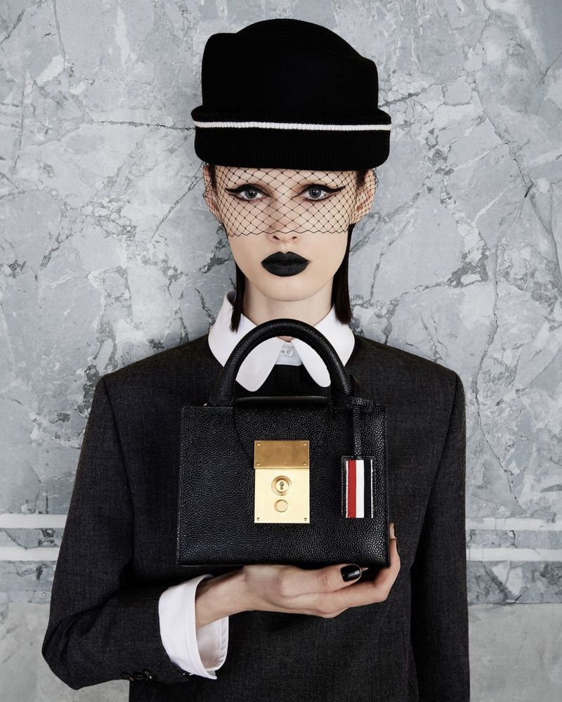 Fashion model with black lipstick and veiled hat holds black leather handbag with gold hardware against a marble background indoors.