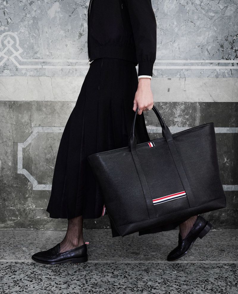 Woman in a black outfit carries a large black leather tote bag with red, white, and blue stripes on a mottled background.