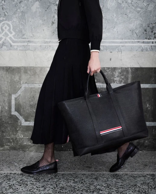 Woman in a black outfit carries a large black leather tote bag with red, white, and blue stripes on a mottled background.