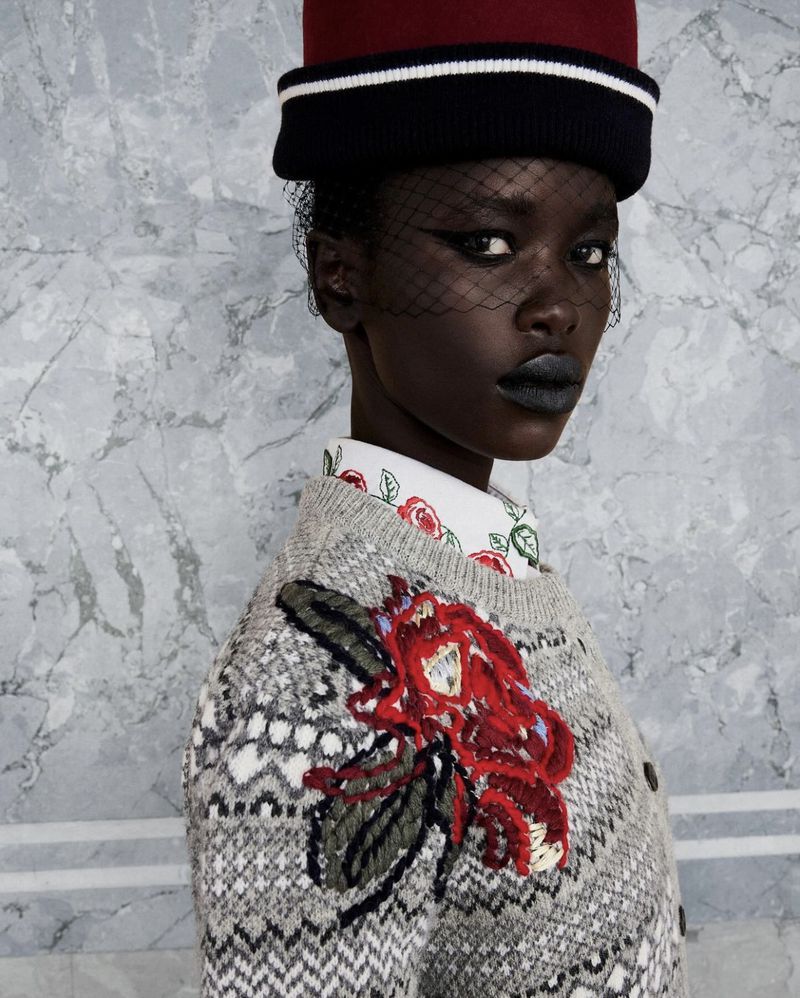 Close-up of a Black fashion model wearing a red pillbox hat, black veil, grey floral knit sweater against a grey marble backdrop.