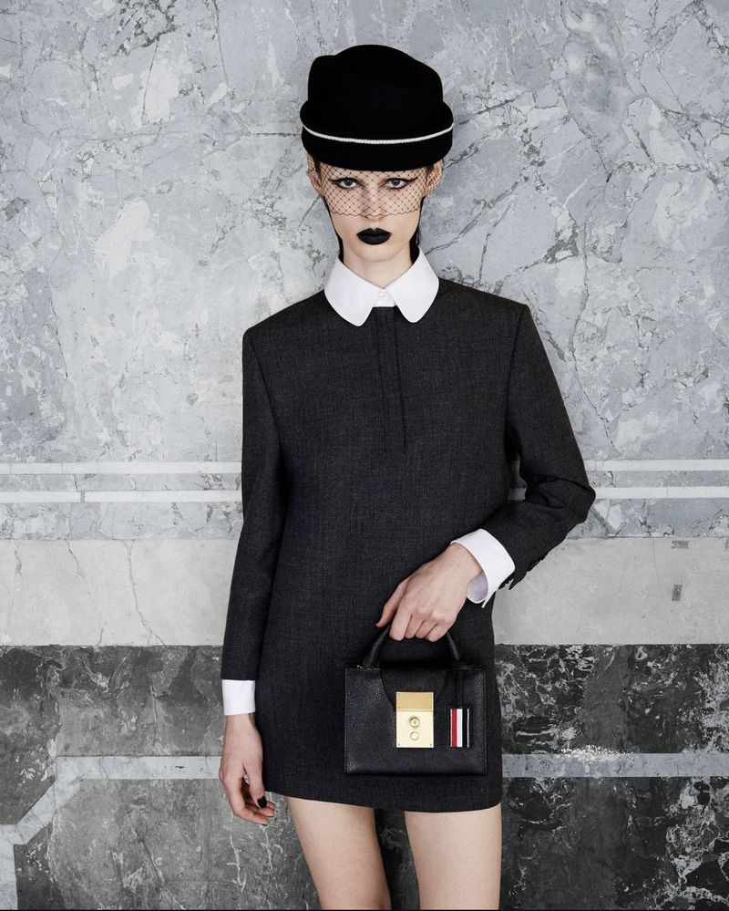 Fashion model poses in a gray dress with white collar, black hat, and netting, holding a structured black leather handbag against a marble wall.