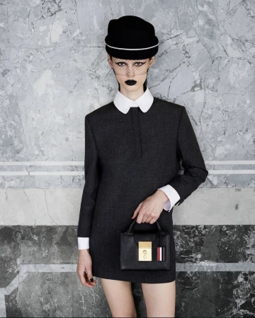 Fashion model poses in a gray dress with white collar, black hat, and netting, holding a structured black leather handbag against a marble wall.