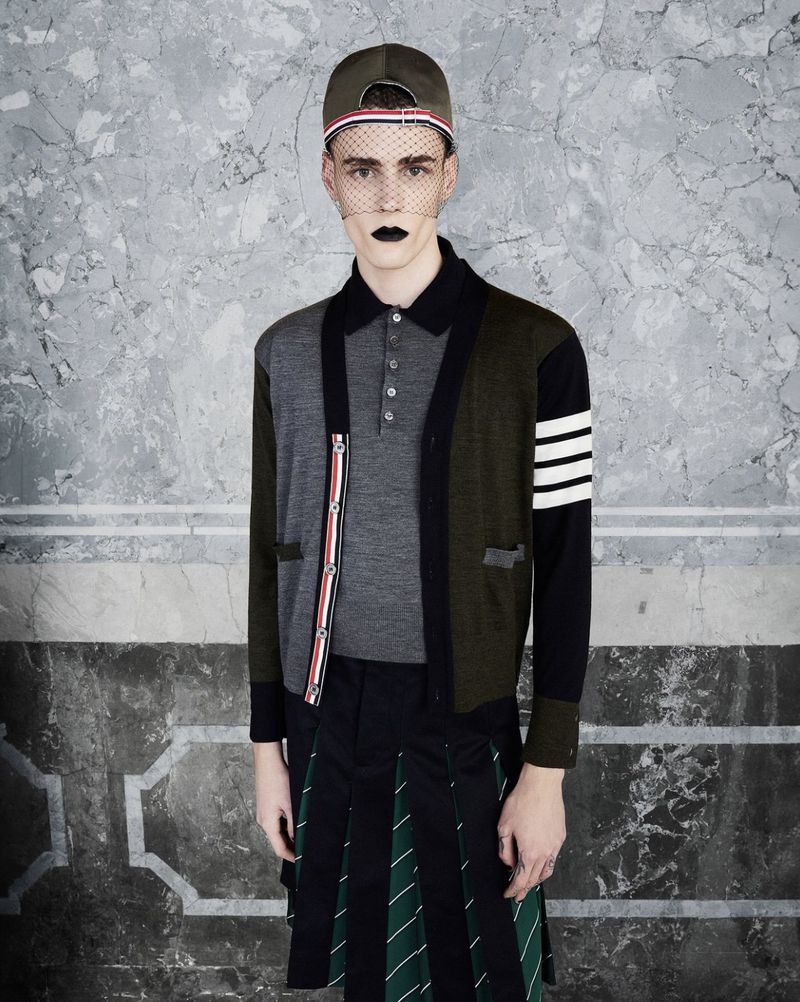 Male fashion model in a gray polo shirt, patchwork cardigan, and kilt-style skirt poses against a textured grey studio backdrop.