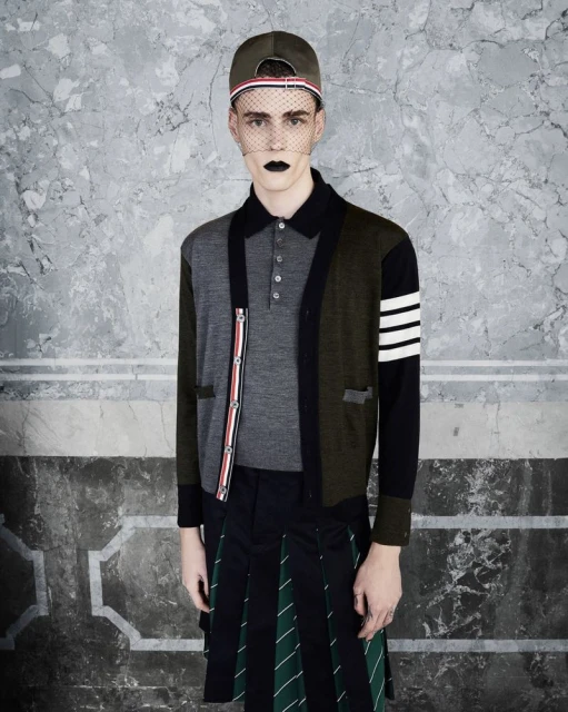 Male fashion model in a gray polo shirt, patchwork cardigan, and kilt-style skirt poses against a textured grey studio backdrop.
