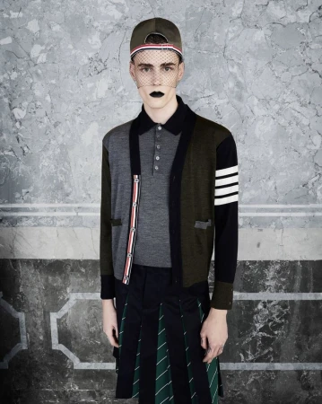 Male fashion model in a gray polo shirt, patchwork cardigan, and kilt-style skirt poses against a textured grey studio backdrop.