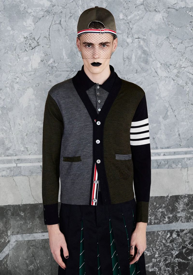 Male fashion model poses in Thom Browne colorblock cardigan, black pleated skirt, and baseball cap against a marbled backdrop.