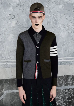 Male fashion model poses in Thom Browne colorblock cardigan, black pleated skirt, and baseball cap against a marbled backdrop.