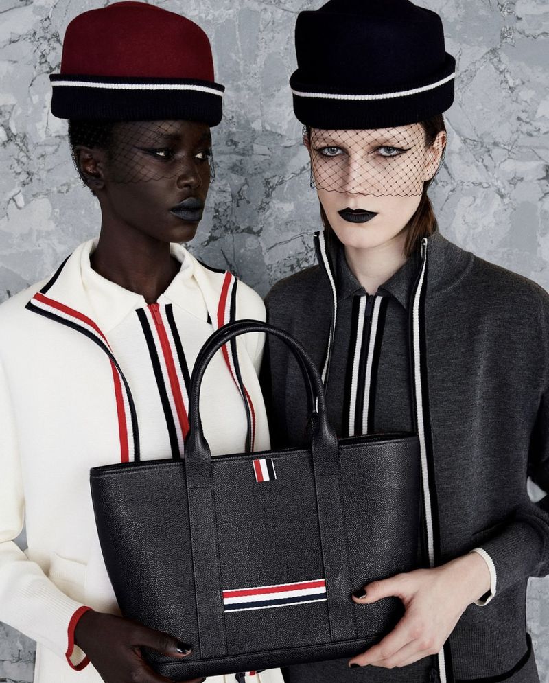 Two fashion models pose in front of a grey mottled wall wearing hats and fashionable outfits while holding a black leather tote.