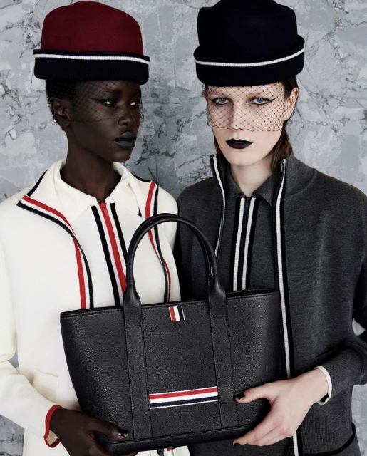 Two fashion models pose in front of a grey mottled wall wearing hats and fashionable outfits while holding a black leather tote.