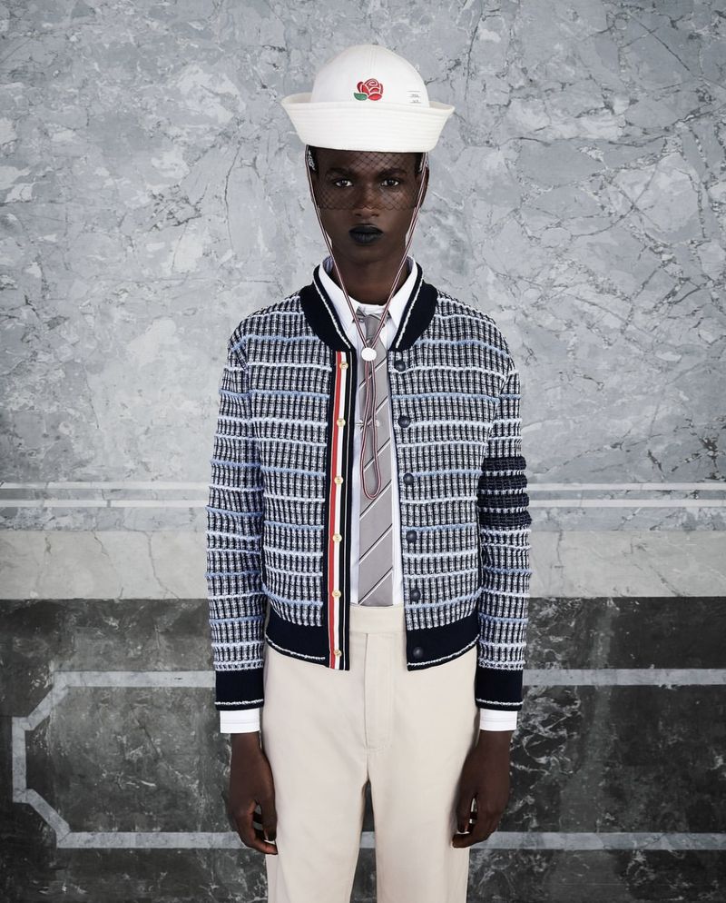 Black model wearing a white sailor hat, patterned bomber jacket, tie, and light trousers against marble wall