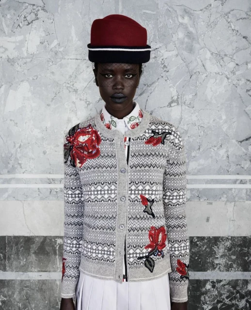 Elegant woman in a gray patterned cardigan with red rose embellishments, wearing a red hat, and standing in front of a marble wall.