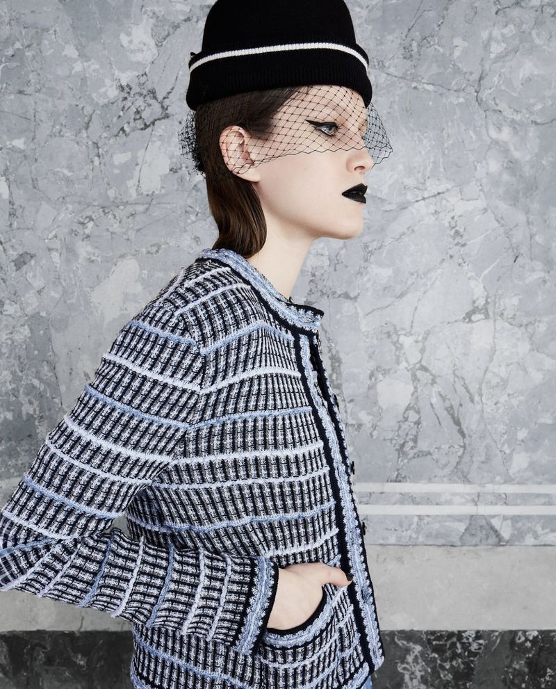 Fashionable woman in jacket and black hat with netting poses against marble background in a studio shot showcasing an elegant look.