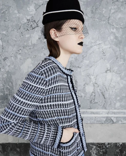 Fashionable woman in jacket and black hat with netting poses against marble background in a studio shot showcasing an elegant look.