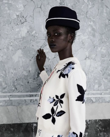 Black model wearing an embroidered white cardigan and dark hat with a veil against a grey marble wall.