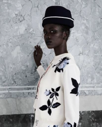 Black model wearing an embroidered white cardigan and dark hat with a veil against a grey marble wall.