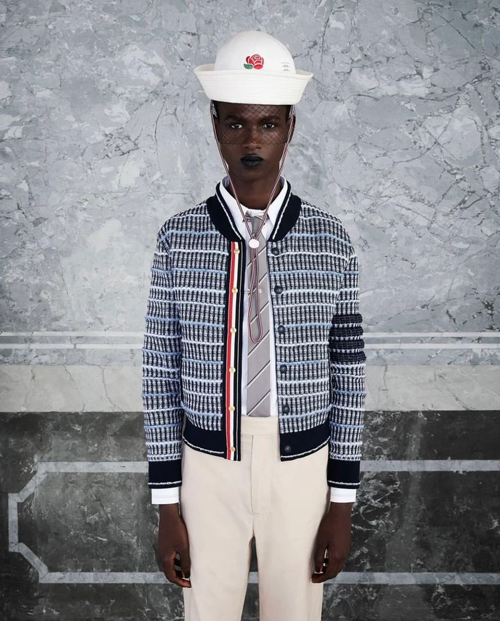 Black model wearing a white sailor hat, patterned bomber jacket, tie, and light trousers against marble wall
