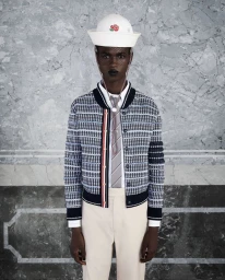 Black model wearing a white sailor hat, patterned bomber jacket, tie, and light trousers against marble wall