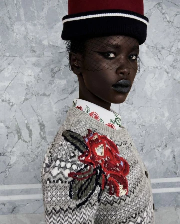 Model wearing a patterned knit sweater with floral embroidery and a red-and-black pillbox hat with birdcage veil.