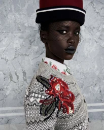 Model wearing a patterned knit sweater with floral embroidery and a red-and-black pillbox hat with birdcage veil.