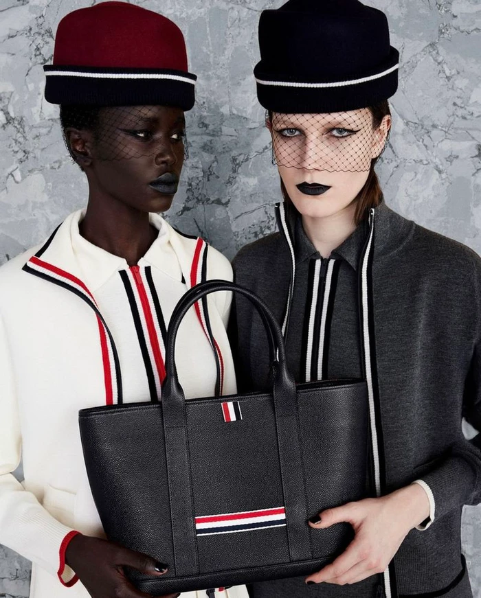 Two models wearing structured hats and veils holding a black leather tote bag for Thom Browne F/W 24
