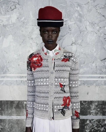 Model wearing a grey patterned cardigan with red floral appliqués and a structured red hat against a veined marble wall.