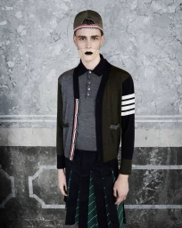 Model wearing a cardigan, pleated skirt, cap with veil, and dark lipstick against a textured grey wall.