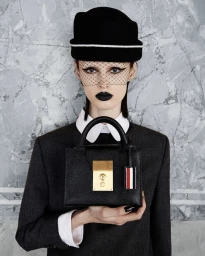 Model wearing a dark cap with net veil and black lipstick holding a structured leather handbag for Thom Browne F/W 24