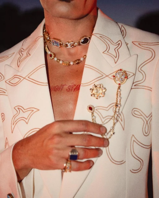 Close-up of a person in a white jacket with gold jewelry including necklaces and rings, adorned with gemstones and pearls.