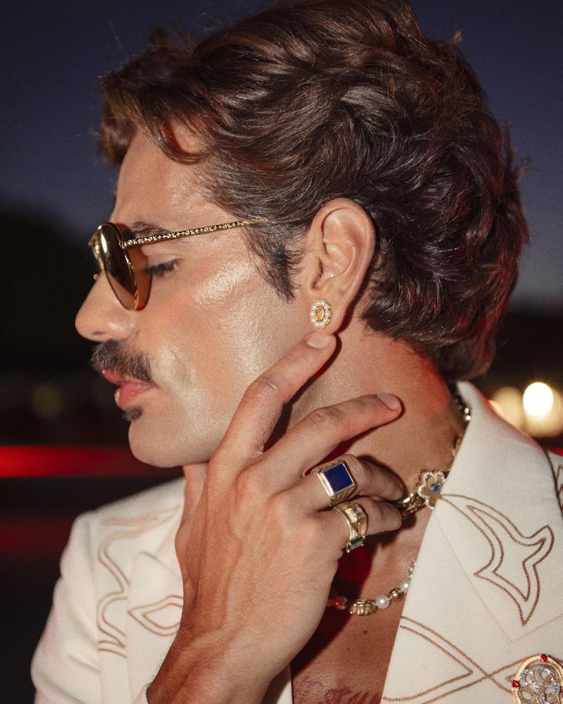 Close-up of adult male with mustache, wearing sunglasses, earring, rings, and white jacket with decorative trim against dark background.