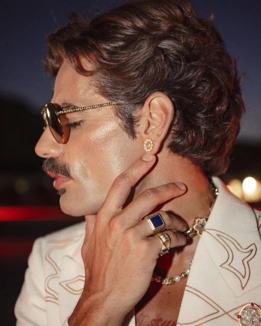 Close-up of adult male with mustache, wearing sunglasses, earring, rings, and white jacket with decorative trim against dark background.