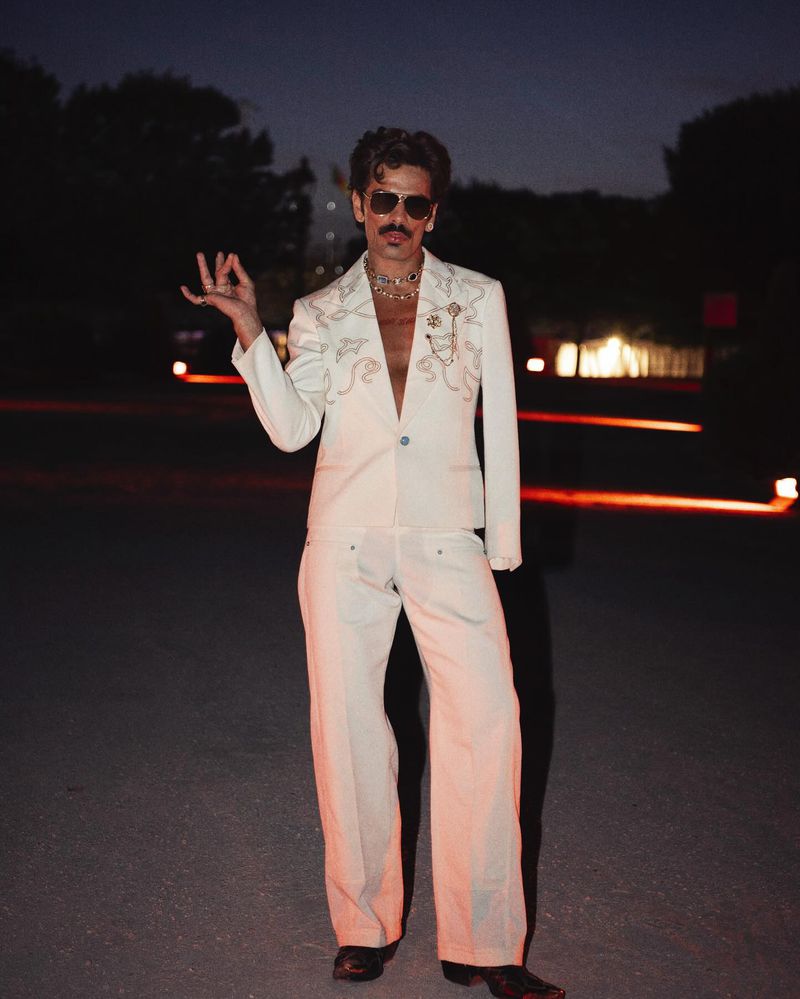Adult male in a white suit with a mustache strikes a pose in an outdoor nighttime setting with trees and building lights in the background.