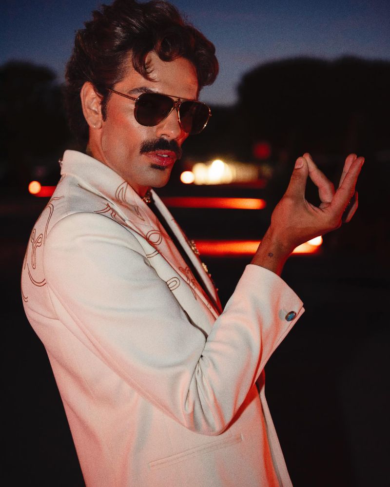 Man in white suit and sunglasses poses outside at night. The stylish adult male has a mustache and dark hair.