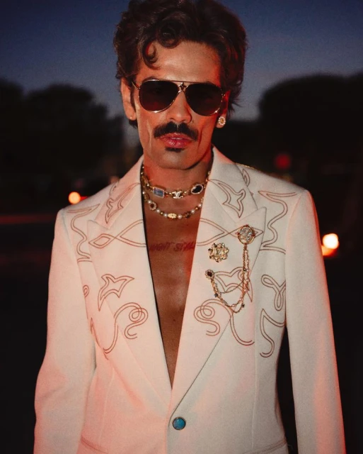 Man in white patterned suit with dark sunglasses, gold necklaces, and a mustache, against a dark outdoor background.