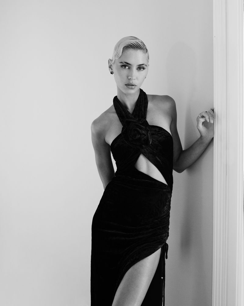 Black-and-white portrait of a woman in a black velvet halter dress posing against a white wall, highlighting fashion and portrait photography.