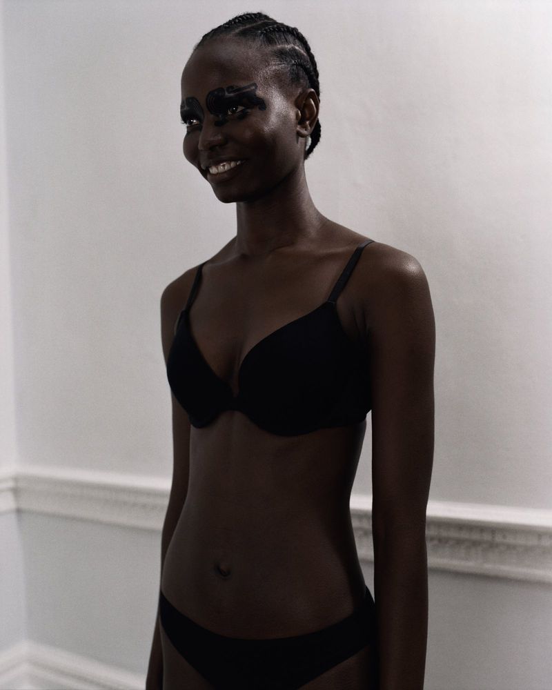 Smiling dark-skinned woman in bra and underwear with eye makeup standing indoors against a plain white wall with molding detail.